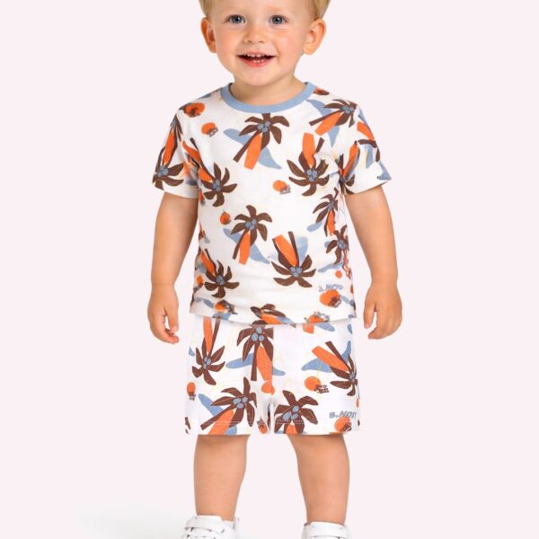 Lachende blonde peuter in zomerpyjama met palmbomenprint en witte sneakers op roze achtergrond