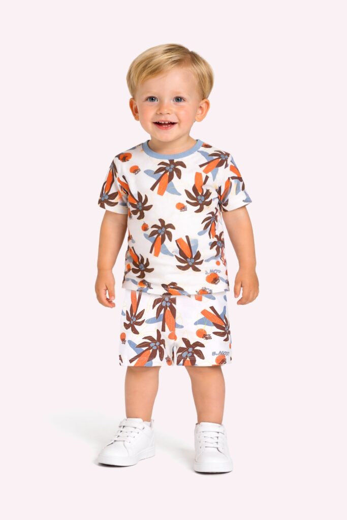 Lachende blonde peuter in zomerpyjama met palmbomenprint en witte sneakers op roze achtergrond