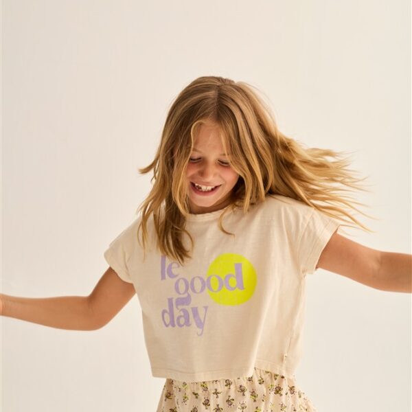Lachend meisje met zwaaiend haar draagt wit T-shirt met tekst good day en gebloemde rok