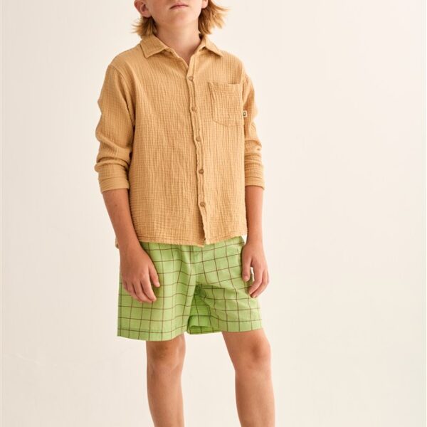 Jongen met beige overhemd, groene geruite short en pet op houten platform tegen witte achtergrond