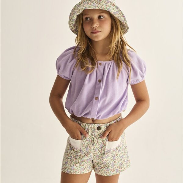 Meisje met bloemenhoedje, lila blouse en gebloemde short met zakken in zomerse outfit