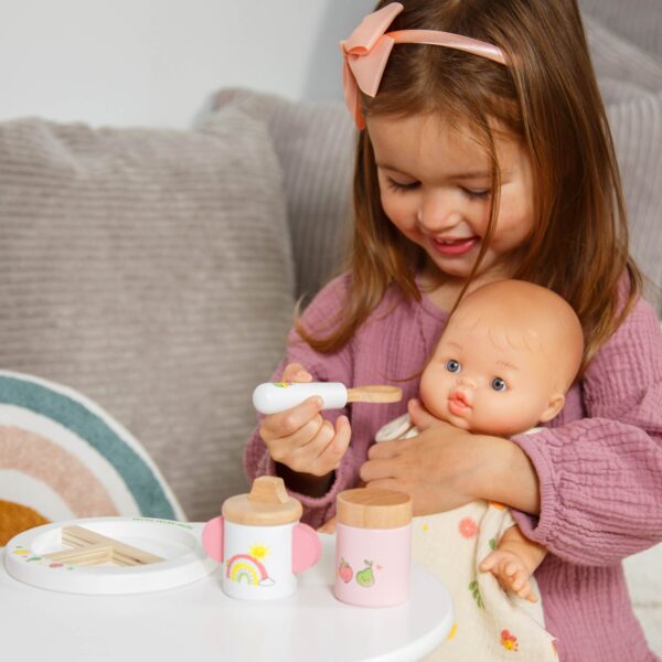 Meisje met roze haarband speelt met babypop en houten speelgoed voedingsset aan tafel